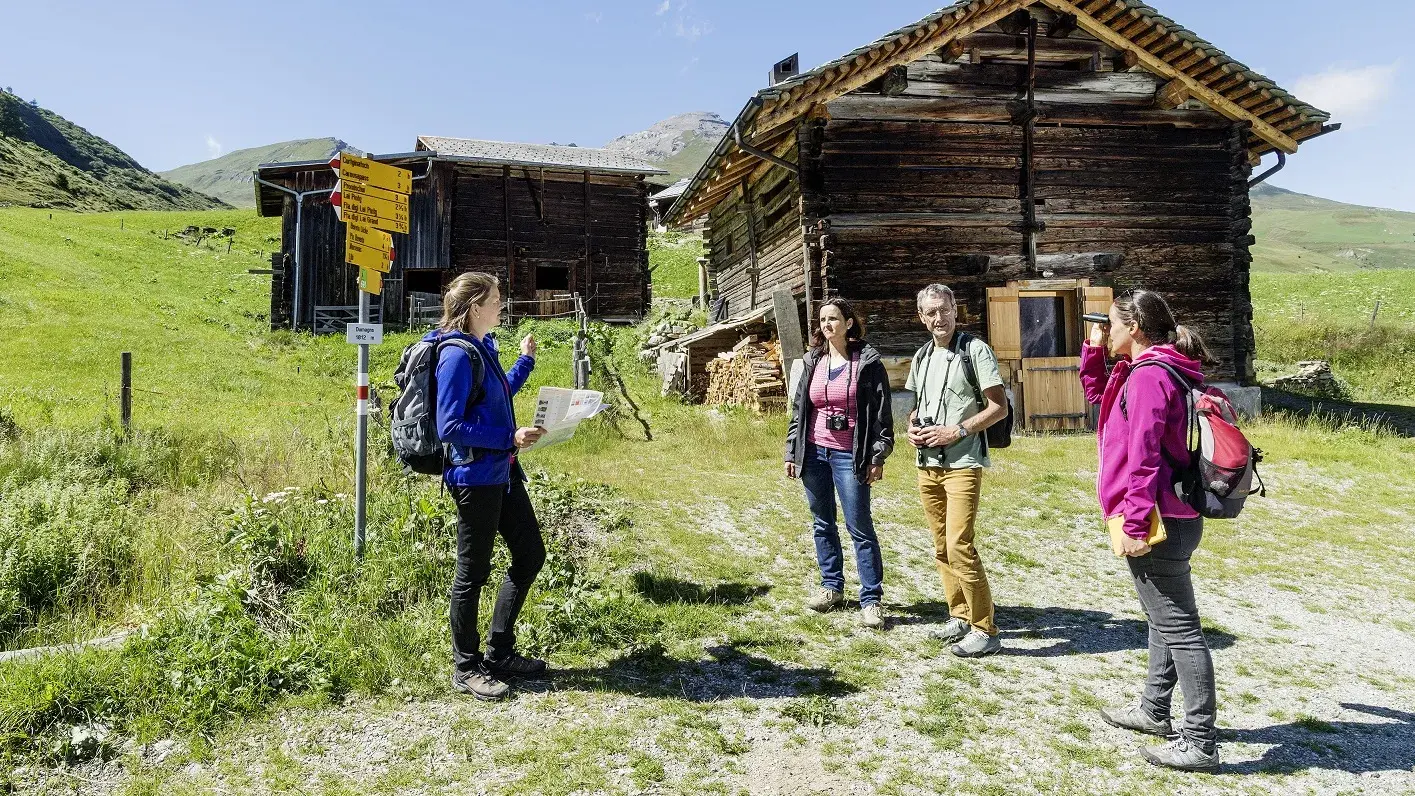 Eine Frau hält einen Vortrag vor einer Gruppe, im Hintergrund sieht man Wanderschilder und Schweizer Alphütten.