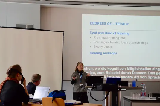 Hörsaal. Daniela Eichmeyer präsentiert ihren Vortrag. Im Hintergrund eine ihrer Vortragsfolien. Sie trägt den Titel: Degrees of Literacy. Deaf and Hard of Hearing.