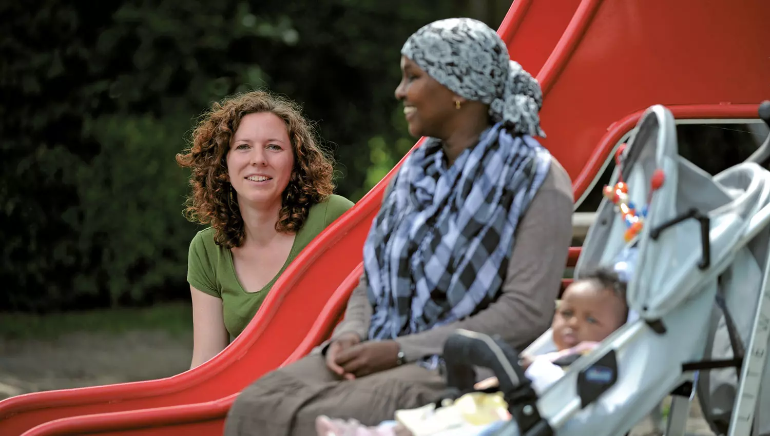 Zwei Frauen sitzen mit Kleinkind auf dem Spielplatz. 