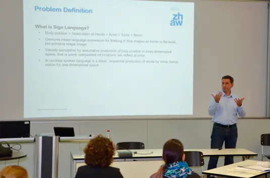 Hörsaal. Andri Reichenbacher präsentiert seinen Vortrag. Im Hintergrund eine seiner Vortragsfolien. Sie trägt den Titel: Problem Definition. What is Sign Language?