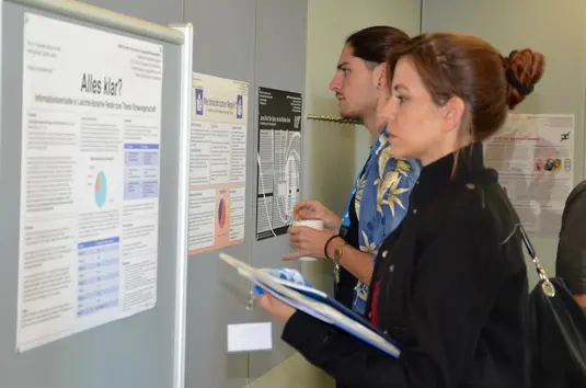 Konferenzbüro. Zwei Teilnehmende stehen vor einer Stellwand mit einem Poster der Poster-Session. Im Hintergrund weitere Poster, die an der Wand des Raums aufgehängt sind.