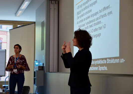 Hörsaal. Irene Strasly trägt ihre Präsentation vor, eine Gebärdendolmetscherin übersetzt, beide Frauen stehen.