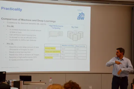 Hörsaal. Andri Reichenbacher präsentiert seinen Vortrag. Im Hintergrund eine seiner Vortragsfolien. Sie trägt den Titel: Practicality. Comparision of Machine and Deep Learnings.