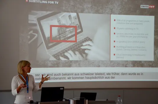 Hörsaal. Michaela Nachtrab präsentiert ihren Vortrag. Im Hintergrund eine ihrer Vortragsfolien mit dem Titel: Subtitling for TV.