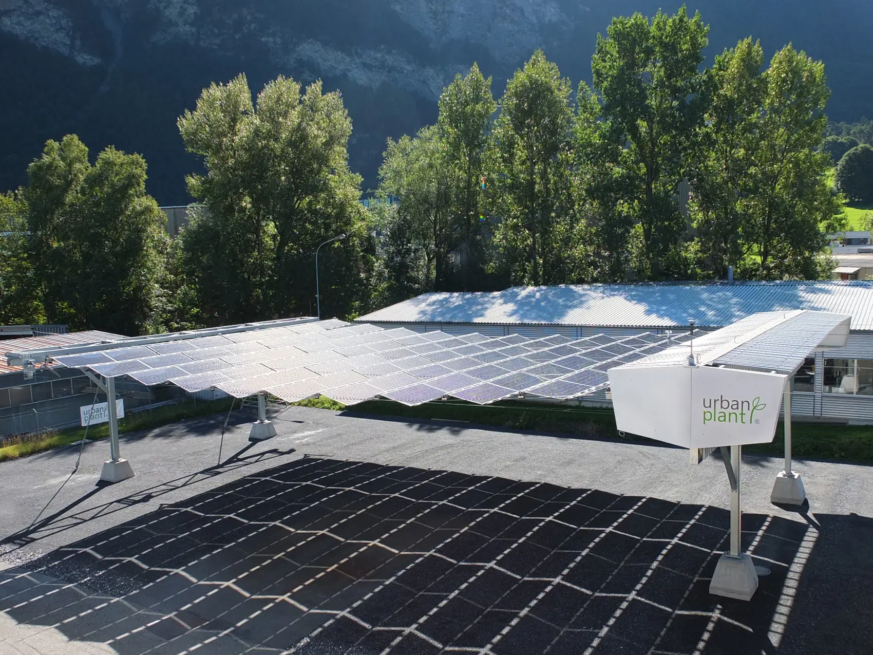 Das Bild zeigt ein Dach aus gefalteten Solarmodulen, das einen grossen Schatten auf den darunterliegenden Parkplatz wirft. Man erkennt das Logo von Urban Plant