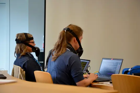 Hörsaal. Rückansicht der beiden Schriftdolmetscherinnen während der Arbeit. Sie tragen Stenomasken und haben ihre Laptops vor sich auf dem Tisch.